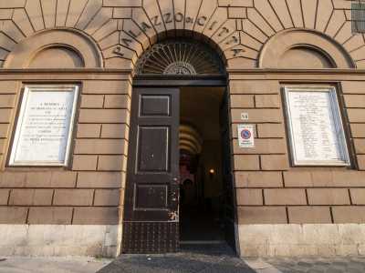 L�iscrizione in lettere di bronzo sormonta la lunetta del portone, la quale si apre nella fascia in bugnato color terra della struttura